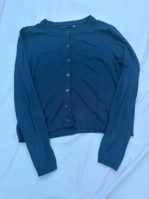 Mörkblå cardigan med knappar - En stilren mörkblå cardigan med rund halsringning och knäppning framtill. Långa ärmar och ribbade muddar vid ärmslut och nederkant. Perfekt att slänga över en t-shirt eller klänning för en enkel layering.
