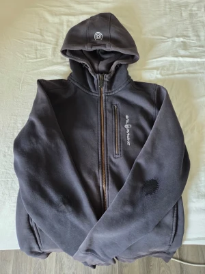 Svart hoodie från Sail Racing - Svart hoodie med dragkedja från Sail Racing. Hoodien har broderad logga på bröstet, fickor med dragkedja och en stor huva med tryckt märke. Tillverkad i mjukt material, perfekt för en avslappnad stil.