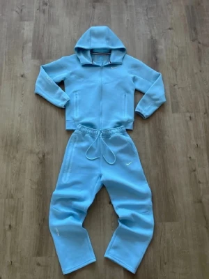 Nike NOCTA ljusblå loungewear set - Säljer ett ljusblått loungewear set från Nike NOCTA. Setet består av en hoodie med dragkedja och matchande byxor med resår och dragsko i midjan. Båda delarna har diskreta Nike-loggor. Den har oljefläk och den behöver tvättas på grund av doft. Perfekt för chill dagar hemma eller när du vill vara bekväm.