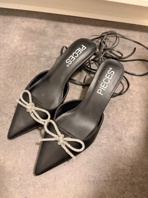 Svarta pumps med strassrosett från PIECES - Snygga svarta pumps från PIECES med spetsig tå och dekorativ strassrosett framtill. Skorna har tunna remmar som knyts runt ankeln för en trendig look. Materialet är konstläder och de har en elegant, feminin siluett.