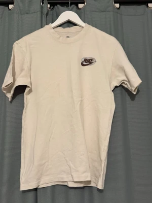 Beige Nike t-shirt med tryck - Säljer en beige t-shirt från Nike med liten logga på bröstet och tryck på ryggen med texten 'PLAY IT COOL FOR MOTHER EARTH'. Klassisk passform och rund halsringning. Perfekt för dig som gillar streetwear och vill ha en clean look.