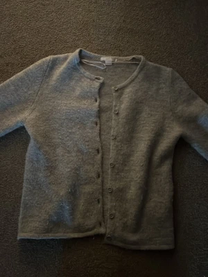 Ljusgrå kofta från Gina tricot - Mysig ljusgrå kofta med rund halsringning och knäppning framtill. Koftan har lång ärm och är tillverkad i ett mjukt, luddigt material som ger en avslappnad look. Perfekt att slänga över en topp för en enkel och stilren outfit.