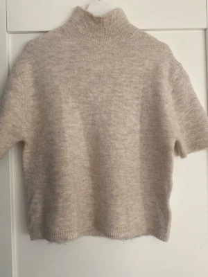 Beige stickad t-shirt med polokrage - Mysig beige stickad t-shirt med korta ärmar och hög polokrage. Tröjan har en mjuk, fluffig känsla och är ribbstickad vid halsen och nederkanten. Perfekt för lager på lager och en stilren look.
