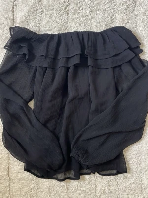 Svart off shoulder blus med volang - Trendig svart off shoulder-blus med volang upptill och långa, luftiga ärmar. Blusen har en lätt transparent känsla och är perfekt för en stilren look. Passar dig som gillar en loose fit och vill ha något snyggt och bekvämt.