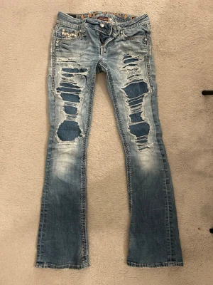 Blå slitna bootcut-jeans från Rock Revival - Säljer ett par ljusblå lowwaist bootcut-jeans från Rock Revival med kraftiga slitningar och patchade hål framtill. Jeansen har dekorativa bakfickor med broderier och detaljer i silver. Tillverkade i denim med klassisk femficksdesign och låg midja. Har haft några slitningar och hål men allt är lagat. Storlek 25. Midjemått 35cm, innerbenslängd 75cm. 