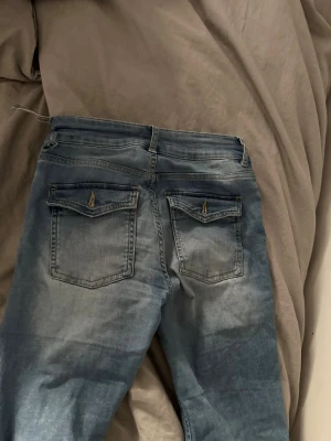Ljusblå slitna jeans med hål - Säljer ett par ljusblå jeans med slitningar och stora hål vid knäna. Jeansen har  två bakfickor med lock och knapp samt klassisk femficksdesign. Perfekta för en avslappnad och trendig look. Dem har ett liket hål på sidan som säkert går att fixa 