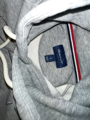 Grå hoodie från GANT med logga - Snygg grå hoodie från GANT med broderad logga och amerikanskt inspirerat märke på bröstet. Hoodien har huva med vita snören och en stor magficka. Perfekt för en avslappnad och sportig stil.