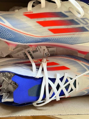 Adidas X Speedflow.1 FG fotbollsskor F50 PROS - F50 pros. Snabba Adidas X Speedflow.1 FG fotbollsskor i vitt med röda och blå detaljer. Skorna har en lätt ovandel i syntetmaterial med FiberTouch-teknologi och klassiska tre ränder på sidan. Designade för spel på naturgräs och ger grym känsla och kontroll på planen.