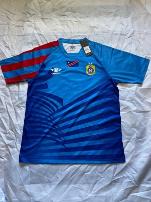 Blå fotbollströja DR Kongo Umbro - Snygg blå fotbollströja från Umbro med DR Kongos flagga och klubbmärke på bröstet. Tröjan har röda och blå ränder på ena ärmen och coolt grafiskt mönster framtill. Perfekt för dig som gillar fotboll och vill sticka ut på planen.