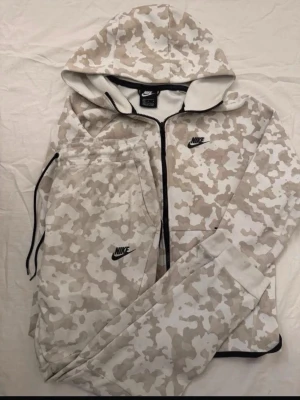 Nike mjukisbyxor camo beige/vit - Nike mjukisbyxor i beige och vit camo-mönster med svart Nike-logga på benet. Byxorna har dragsko i midjan och är gjorda i mjukt material för maximal komfort. Perfekt för chill eller träning, matchar med hoodie i samma stil.