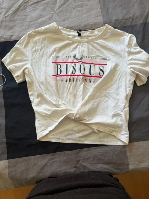 Vit croppad t-shirt med tryck - Säljer en vit croppad t-shirt med trycket 'BISOUS PARISIENNE' i svart och rött framtill. T-shirten har korta ärmar och en knutdetalj längst ner. Perfekt för dig som gillar en avslappnad och trendig stil.