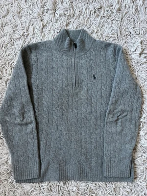 Polo ralph lauren grå cableknit - Mysig grå stickad tröja med hög polokrage och ribbade muddar. Perfekt för kyliga dagar och enkel att matcha med jeans eller kjol. Tröjan har en klassisk rak passform och är gjort på kashmir och ull.