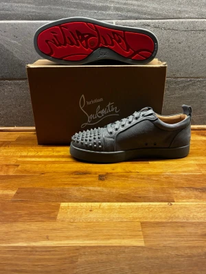 Grå Christian Louboutin sneakers - Snygga grå sneakers från Christian Louboutin med ikoniska röda sulor och nitar på tån. Skorna har lågt skaft, snörning och är tillverkade i läder. Perfekta för dig som vill sticka ut med en exklusiv och edgy look.