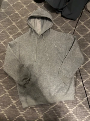 Grå hoodie med broderad logga - Snygg grå hoodie med broderad vit logga på bröstet. Hoodien har en klassisk huva med dragsko och en stor magficka. Tillverkad i mjukt och skönt material med borstad insida för extra komfort. Perfekt för en avslappnad och trendig look.