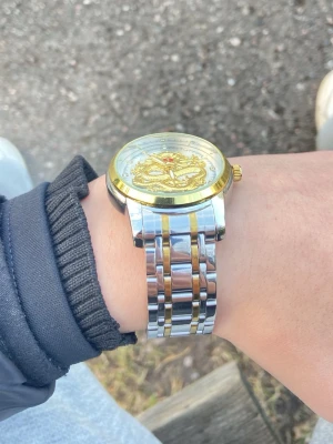 Silver och guld klocka med drakmotiv - Stilren klocka i silver och guld med ett detaljerat drakmotiv på urtavlan. Armbandet är i metall med både silver- och guldfärgade länkar. Urtavlan har gulddetaljer och en unik design som sticker ut.