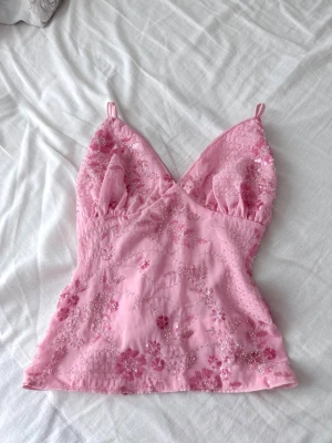 Rosa blommig linnetopp med paljetter - Supersöt rosa linnetopp med tunna axelband och v-ringning. Toppen har blommiga broderier och paljettdetaljer som ger ett glittrigt intryck. Perfekt för dig som gillar romantisk och feminin stil.