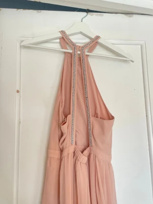 Lång ljusrosa halterneckklänning - Elegant långklänning i ljusrosa med halterneck och glittriga detaljer längs halsringningen. Klänningen har plisserad kjol och öppen rygg. Bra som examen, bal eller finklänning. Går inte att köpa längre