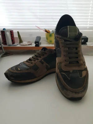 Valentino Camo Rockrunner - Snygga sneakers från Valentino med coolt camo-mönster i grönt, brunt och svart. Skorna har detaljer i mocka och läder, samt klassisk låg profil med snörning. Perfekta för dig som vill sticka ut med exklusiva och trendiga sneakers.