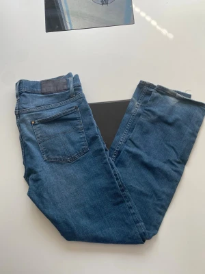 Blå raka jeans i klassisk modell - Säljer ett par blå jeans i klassisk rak modell med fem fickor och bälteshällor. Jeansen har en medelhög midja och är tillverkade i denim. Perfekta för en avslappnad och stilren look.
