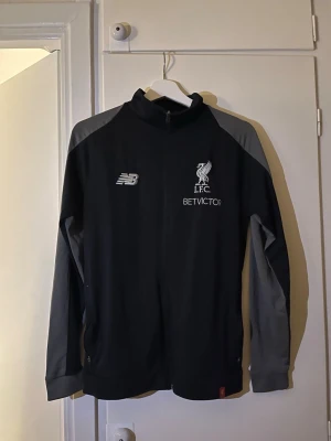 Liverpool träningsjacka New Balance - Snygg svart och grå träningsjacka från New Balance med Liverpool FC-logga och BETVICTOR-tryck på bröstet. Jackan har hel dragkedja, ståkrage och långa ärmar. Perfekt för fotbollsträning eller som sportig vardagsstil.