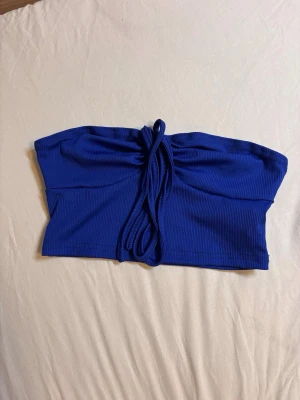 Blå ribbad bandeau tubtopp - Snygg blå bandeau tubtopp med ribbad struktur och knytband framtill. Toppen är ärmlös och har en croppad passform, perfekt till sommarens outfits.