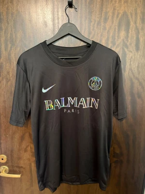 Svart PSG x Balmain t-shirt Nike - Snygg svart t-shirt från Nike i samarbete med PSG och Balmain. Tröjan har holografiska loggor och text, rund halsringning och är tillverkad i ett lätt, snabbtorkande Dri-FIT-material. Perfekt för träning eller streetwear.