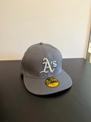 Grå New Era 59FIFTY A's keps - Snygg grå keps från New Era 59FIFTY med Oakland A's logga broderad framtill. Klassisk flat brim och autentisk MLB-stil. Kepsen är i ett stadigt material och har märkets detaljer invändigt. Perfekt för dig som gillar sportig streetstyle.