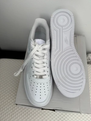 Nike Air Force 1 vita sneakers - Klassiska Nike Air Force 1 sneakers i helvitt läder med perforerad tå, vit sula och ikonisk Swoosh-logga på sidan. Skorna har lågt skaft, vita skosnören och metallbricka med 'AF1' på snörningen. Perfekta för en clean och stilren look. Boxen är lite skadad men skorna är i nytt skick