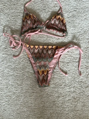 Mönstrad bikini med knyt i sidorna - Säljer en snygg bikini med zigzagmönster i rosa, grön, gul och brun. Både överdel och underdel har knytband i sidorna och runt nacken. Perfekt för sommaren! Har aldrig använd och köpt utomlands på en marknad så vet inte vart den är ifrån 💕