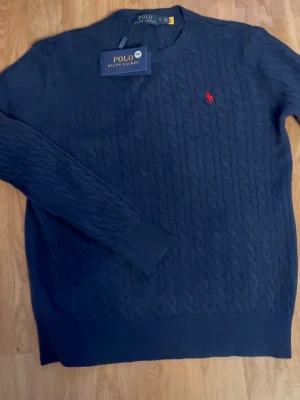 Mörkblå kabelstickad tröja Polo Ralph Lauren - Kabelstickad mörkblå tröja från Polo Ralph Lauren med klassisk röd broderad logga på bröstet. Tröjan har rund halsringning, ribbade muddar och är långärmad. Perfekt för dig som gillar stilren och tidlös design.