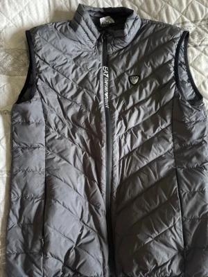 Grå dunväst från Emporio Armani - Snygg grå dunväst från Emporio Armani EA7 med dragkedja framtill och logotyp på bröstet. Västen har ståkrage och diskreta svarta detaljer längs ärmhålen. Perfekt för lager-på-lager och streetwear.