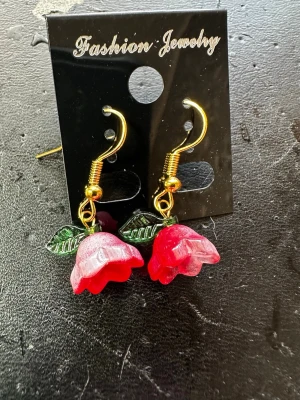 Röda rosörhängen med guldkrok - Snygga örhängen med röda rosor i plast och gröna blad. Krokarna är guldfärgade och ger en cool kontrast till de färgstarka blommorna. Perfekta för dig som vill sticka ut med ett lekfullt och romantiskt smycke.