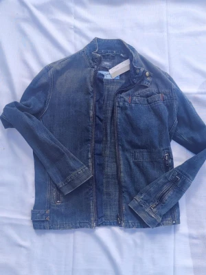 Oanvända G-Star Raw design jeans jacka med etikett  - Oanvänd snygg, blå och cool design jeansjacka från G-Star Raw med ståkrage och dragkedja framtill. Jackan har flera fickor, detaljerade sömmar och justerbara spännen vid ärmslut och midja. Passar perfekt till en avslappnad streetstil och har en modern, slim fit design. Se bilder för prisetikett.