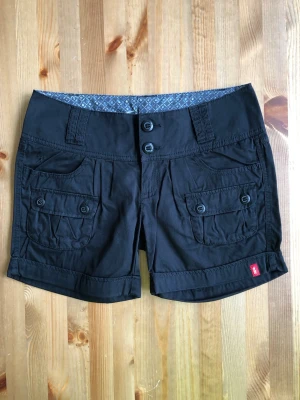 Svarta shorts med fickor från Esprit - Säljer ett par svarta shorts från Esprit med två stora fickor fram och bak, knappar och uppvikta benslut. Shortsen har en midwaist passform och är tillverkade i bomull, perfekta för sommaren.