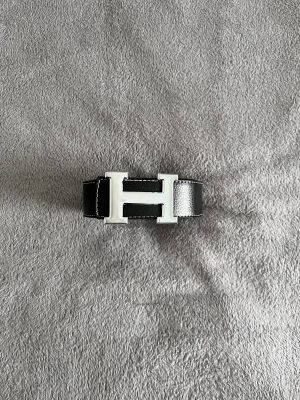 Herme’s bälte  - Stilrent svart skärp med ikoniskt H-format spänne i silverfärg. Skärpet är gjort i slätt skinn och har vita kontrastsömmar längs kanterna. Perfekt accessoar för att lyfta din outfit med en lyxig touch.
