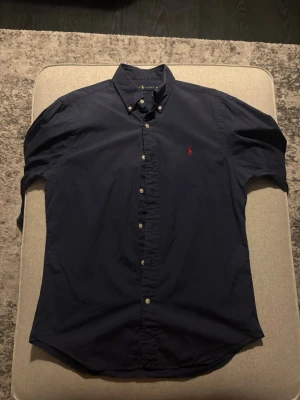 Mörkblå skjorta från Ralph Lauren - Klassisk mörkblå skjorta från Ralph Lauren med button-down krage och röd broderad logga på bröstet. Skjortan har långa ärmar och vita knappar framtill. Perfekt för en stilren och tidlös look.