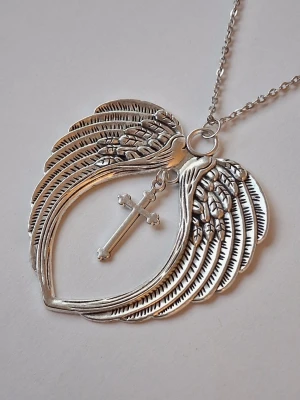 Halsband med änglavingar och kors - Snyggt halsband i silverfärgad metall med ett stort hänge format som två detaljerade änglavingar som bildar ett hjärta. I mitten hänger ett kors. Perfekt accessoar för dig som gillar unika smycken med symbolik.