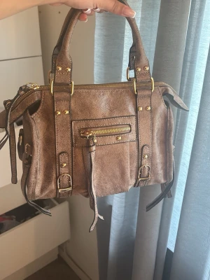 Handväska - Cool brun handväska i mocka med guldfärgade och dragkedjor. Det följer med ett justerbart axelband! Väskans mått: ca 25x18cm. Helt ny, aldrig använt.