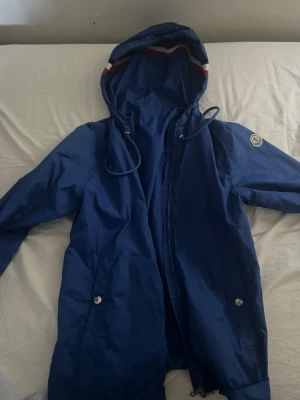 Blå regnjacka från Moncler - Snygg blå regnjacka från Moncler med huva och dragsko. Jackan har vita och röda detaljer på huvan samt Moncler-logga på ärmen. Två fickor med tryckknappar och dragkedja framtill. Perfekt för regniga dagar.