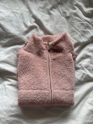 Rosa teddytröja med dragkedja - Mysig rosa teddytröja med hel dragkedja och hög krage. Tröjan är tillverkad i fluffigt, mjukt material och har långa ärmar. Perfekt för kyliga dagar när du vill ha något extra gosigt på dig.