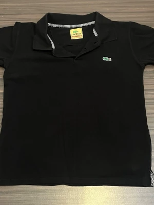Lacoste pikétröja - Klassisk svart pikétröja från Lacoste med korta ärmar och krage. Tröjan har den ikoniska gröna krokodilloggan på bröstet och knäppning vid halsen. Storleken på tröjan är S och skriv till mig innan ni köper den. 