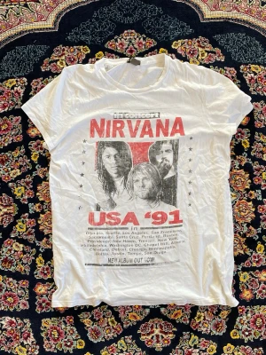 Vit Nirvana t-shirt med tryck - Cool vit t-shirt med stort Nirvana-tryck framtill och texten 'USA '91'. T-shirten har korta ärmar och klassisk passform. Perfekt för dig som gillar bandtröjor och vill ha en avslappnad stil.