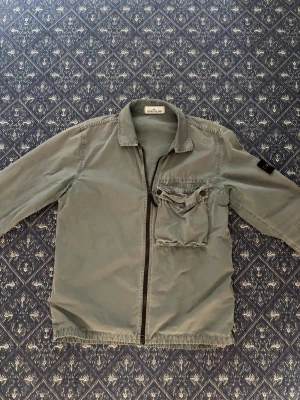 Grå overshirt från Stone Island - Snygg grå overshirt från Stone Island med dragkedja framtill och en stor bröstficka med knapp. Klassisk krage och ikonisk logotyp-patch på ärmen. Perfekt lager-på-lager-plagg i mjukt bomullsmaterial.