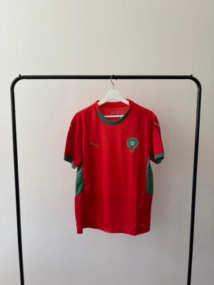 Marocko fotbollströja Puma röd/grön - Säljer en officiell Marocko fotbollströja från Puma i rött med gröna detaljer. Tröjan har korta ärmar, grönt runt halsen och ärmsluten samt Marockos landslagsemblem på bröstet. Tillverkad i lätt och ventilerande material, perfekt för träning eller match.