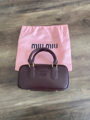 Miu Miu handväska  - Snygg brun handväska från Miu Miu i slät läderlook med guldiga detaljer och dubbla handtag. Väskan har en rektangulär form och logotypen är präglad framtill. Kommer med en rosa dustbag med Miu Miu-tryck.