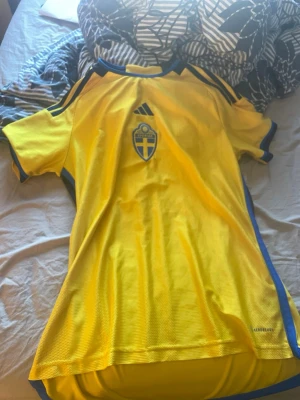 Svensk landslagströja Adidas gul - Gul svensk landslagströja från Adidas med blå detaljer och landslagets emblem på bröstet. Tröjan har korta ärmar, rund hals och är tillverkad i ett ventilerande funktionsmaterial. Perfekt för fotboll eller träning.