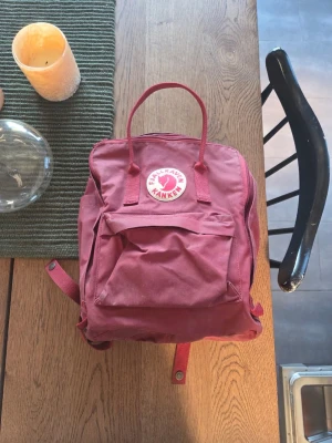 Vinröd Kånken ryggsäck från Fjällräven - Klassisk vinröd Kånken ryggsäck från Fjällräven med ikonisk rund logotyp framtill. Blekt på vissa ställen och behöver en liten tvätt. I övrigt i fint skick! 