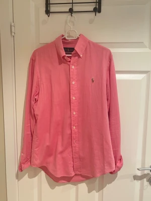 Rosa linne skjorta från Ralph Lauren - Klassisk rosa linne skjorta från Ralph Lauren med button-down krage och broderad logga på bröstet. Skjortan har långa ärmar och knäppning framtill. Perfekt för en stilren och avslappnad look.