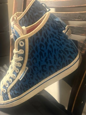 Adidas blå leopardmönstrade sneakers - Unika Adidas sneakers med blått leopardmönster och beige detaljer. Skorna har högt skaft, vita snören och en klassisk Adidas-logga på sidan. Insidan är röd och materialet är canvas. Perfekta för dig som vill sticka ut med din stil.
