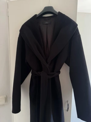Svart kappa med knytskärp från Ellos - Stilren svart kappa från Ellos med bred krage och knytskärp i midjan. Kappan har en rak passform och långa ärmar, perfekt för en trendig och enkel look. Materialet ser ut att vara mjukt och följsamt, vilket ger en snygg siluett.